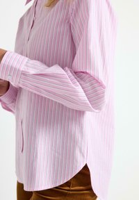 Lyserød stribedeskjorte med tynde lodrette striber i hvid og blå, knaplukning ved manchetten, buet kant og en glat bomuldsstruktur.