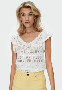 Top de punto blanco con mangas cortas y acampanadas, que presenta un patrón de tejido abierto y un diseño de escote en V. Combinado con pantalones amarillos de talle alto.