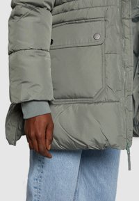 Chaqueta acolchada gris con un bolsillo prominente y detalles de botón a presión. Cuenta con puños acanalados y una textura suave y brillante.