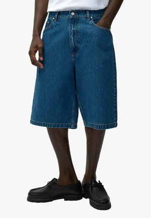 Uomo che indossa pantaloncini blu in denim larghi fino al ginocchio, camicia bianca e scarpe di pelle nere, in piedi con una mano in tasca.