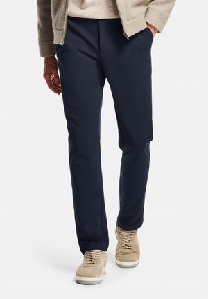 Mann trägt marineblaue Hose, beige Jacke und beige Turnschuhe, steht mit einer Hand in der Tasche vor einem weißen Hintergrund.