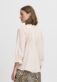 Blusa rosa chiaro con maniche a sbuffo, polsini arricciati e una texture liscia, abbinata a una gonna con stampa leopardata. Vista posteriore mostrata.