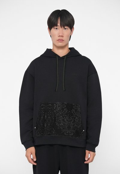 Sort hoodie lavet af blødt stof, der har et tekstureret sort blondermønster på frontlommen og kontrasterende grøn snor. Simpelt design.