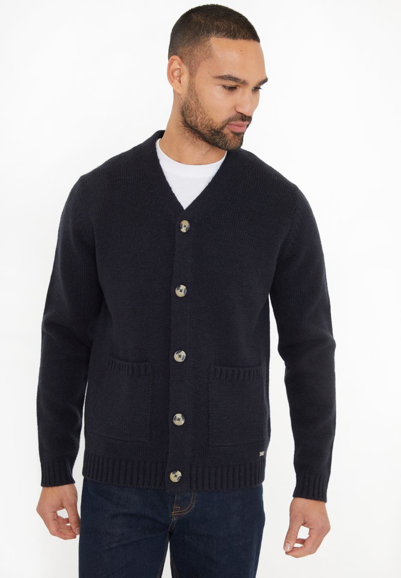 Man in een zwarte gebuttonde cardigan met twee voorzakken over een wit shirt en donkere jeans, kijkend opzij.