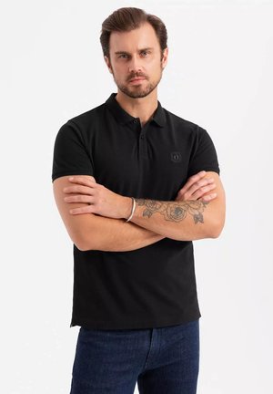 Camisa polo negra de manga corta con cuello, que presenta un logo sutil en el pecho. Está hecha de una tela suave, tiene un diseño ajustado y un dobladillo limpio.