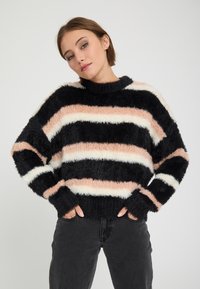 Jeune femme portant un pull rayé duveteux avec des bandes noires, roses et blanches et un pantalon foncé, debout les mains dans les poches.