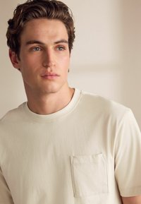 Helle beige Baumwoll-T-Shirt mit rundem Halsausschnitt und einer Brusttasche auf der linken Seite. Weicher Stoff mit lässiger Passform.
