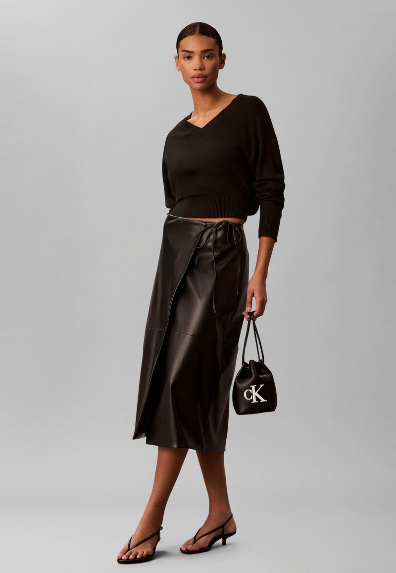 Γυναίκα που φοράει μαύρο cropped top με μακρύ μανίκι, μαύρη δερμάτινη midi φούστα τύπου wrap, κρατάει μικρή μαύρη τσάντα και φοράει μαύρα πέδιλα με τακούνι.