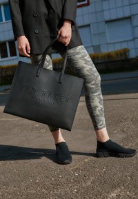 Osoba w kamuflażowych legginsach i czarnych butach trzyma dużą czarną torbę na ramię Ted Baker London na tle miejskim.