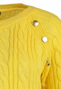 Pull jaune en maille torsadée avec col rond et trois boutons-pression argentés le long de la couture d'épaule.