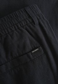 Temne bombažne hlače z elastičnim pasom, ki se ponašajo z gladko teksturo in majhno črno oznako "GABBA" na zadnjem žepu.