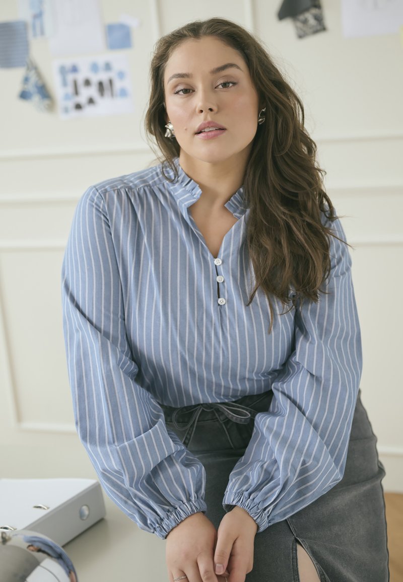 Helles blau gestreiftes Blusenshirt mit rüschigem Ausschnitt, Knopfverschluss und elastischen Bündchen, kombiniert mit einem dunkelgrauen Rock mit einem seitlichen Schlitz.