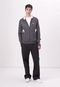 Graue Zip-Jacke mit strukturiertem Design, weißen Akzenten und gerippten Bündchen, kombiniert mit schwarzen weit geschnittenen Hosen und weißen Turnschuhen.