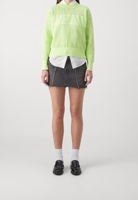 Maglione a maglia verde lime con texture perforata e righe orizzontali, indossato sopra una camicia bianca con bottoni. Gonna mini in denim nera, calze alla caviglia e mocassini.