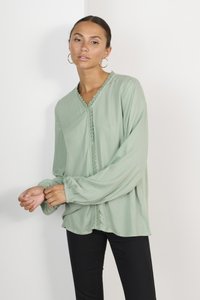Blouse vert clair à manches longues, avec un bord en dentelle le long de l'encolure et du plastron, coupe décontractée et poignets froncés. Associée à un pantalon noir.