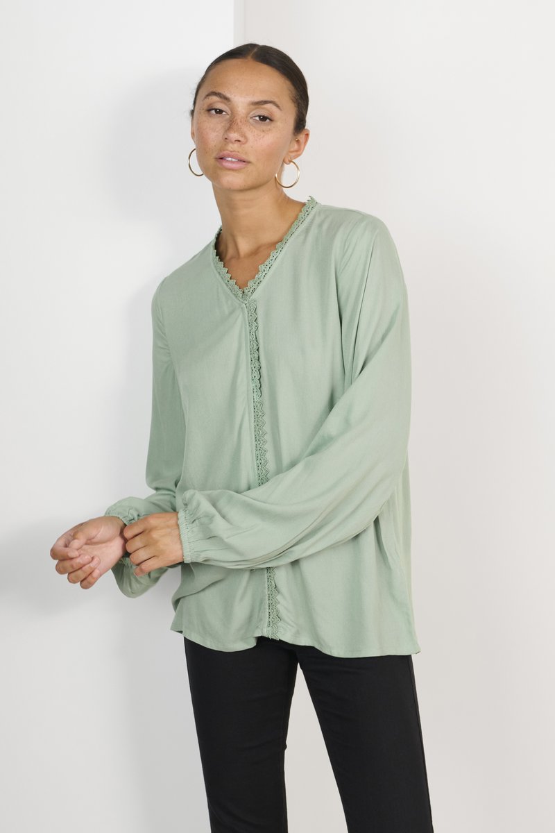 Blouse vert clair à manches longues, avec un bord en dentelle le long de l'encolure et du plastron, coupe décontractée et poignets froncés. Associée à un pantalon noir.