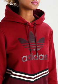 Mörkröd hoodie med en stor svart Adidas-logotyp och tre horisontella ränder i svart och vitt vid fållen. Tillverkad av mjukt tyg med dragsko i huvan.