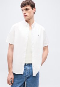 Νέος άνδρας με λευκό πουκάμισο με κοντά μανίκια πάνω από λευκή φανέλα και μπλε τζιν, κοιτάζοντας προς το πλάι.