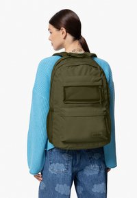Zaino verde oliva con molteplici tasche, dettagli in rete e un tessuto resistente. Il design include spallacci regolabili e una toppa con il logo.