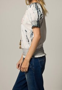 Bílý top s krátkým rukávem a modrými tie-dye akcenty, vyšívaným výstřihem a detailními manžetami. Vytvořeno s modrými džíny; pohodlný střih.