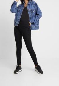 Chaqueta vaquera azul de corte relajado, body negro, leggings negros de talle alto y zapatillas deportivas negras con detalles blancos.