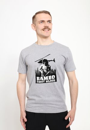 Šedé bavlněné tričko s černou grafikou svalnaté postavy a helikoptérou nad ní, text zní "RAMBO PRVNÍ KRVE" na spodní části.