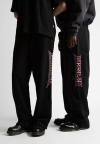Pantalones anchos negros de algodón con texto bordado en rojo que dice "Wasted" en el costado, combinados con zapatos negros y camisetas holgadas.