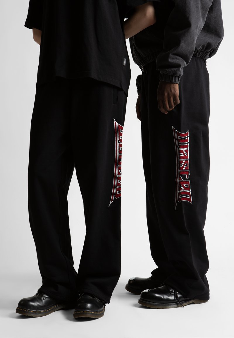 Pantalones anchos negros de algodón con texto bordado en rojo que dice "Wasted" en el costado, combinados con zapatos negros y camisetas holgadas.