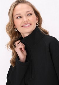 Jachetă neagră cu fermoar, cu guler înalt și textură netedă. Dispune de un fermoar frontal și o croială lejeră, asociată cu cercei rotunzi albi.