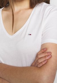 Biała koszulka z krótkim rękawem i dekoltem w serek, z małym haftowanym logo w kolorach czerwonym, białym i niebieskim po lewej stronie klatki piersiowej, wykonana z miękkiej bawełny.
