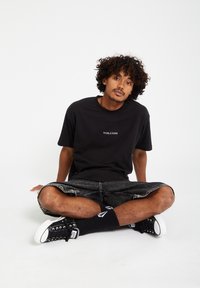 Svart bomull T-skjorte med "VOLCOM"-logoen i hvitt, sammen med mørke denimshorts og svarte sneakers, sitter på en hvit overflate.