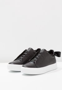 Sneakers in pelle nera con una texture liscia, caratterizzati da un design distintivo, suola in gomma bianca rialzata e un dettaglio a fiocco sul retro.