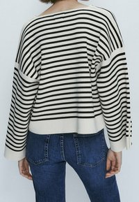 Personne portant un pull ample rayé noir et blanc et un jean bleu, vue de dos sur un fond uni.