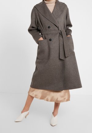 Kobieta ubrana w brązowy kratowany dwurzędowy płaszcz z paskiem, na beżowej satynowej spódnicy i białe mules na obcasie, stojąca z rękami w kieszeniach.