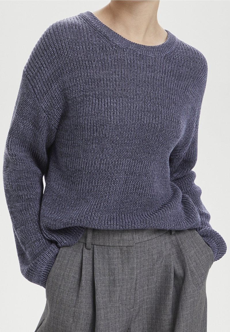 Personne portant un pull en maille bleu marine ample à manches longues, rentré dans un pantalon plissé gris taille haute, les mains dans les poches.