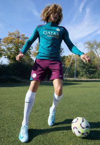Training kit with turquoise long-sleeve top and maroon shorts, featuring logo accents. White knee-high socks and light blue soccer cleats.
