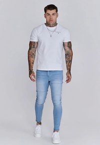 Baltas medvilninis trumpomis rankovėmis marškinėlis su apvalia iškirpte, derinamas su šviesiai mėlynais aptemptais džinsais ir baltais sportbačiais. Matomi tatuiruočių piešiniai ant rankų.