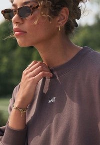 Smeđa majica s kapuljačom s natpisom "self.", okruglim izrezom i patentnim zatvaračima na rukavima. Model nosi sunčane naočale u obliku kornjačevine i zlatnu narukvicu.