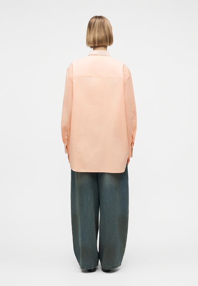 Άτομο με κοντά ξανθά μαλλιά που φοράει ένα μακρύ ροδακινί πουκάμισο και φαρδιά σκούρα γκρι παντελόνια, στέκεται με την πλάτη προς τα εμπρός σε ένα απλό φόντο.