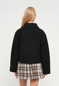 Μαύρο cropped  σακάκι με γιακά με εξάλειψη, μακριά μανίκια και ομαλή υφή, συνδυασμένο με φούστα καρό σε ζεστούς τόνους.