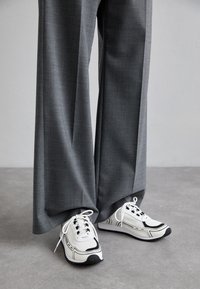 Baskets blanches et noires avec un tissu texturé et une semelle à motifs, associées à un pantalon gris, ample et taille haute.