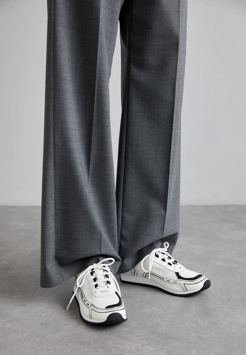 Baskets blanches et noires avec un tissu texturé et une semelle à motifs, associées à un pantalon gris, ample et taille haute.