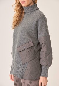 El pulóver gris presenta un cuello alto, texturas mixtas, un panel acolchado con un bolsillo de solapa y acentos acanalados en las mangas y el dobladillo.