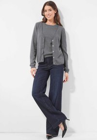 Frau lächelt und lehnt sich an eine Wand, trägt einen grauen Cardigan und Pullover, dunkle weit geschnittene Jeans und schwarze High Heels.