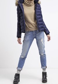 Osoba v námořnické péřové bundě s kožíškem na kapuci, béžovém roláku, ošuntělých džínech s ohrnutými nohavicemi, vzorovaných ponožkách a černých kotníkových botách.