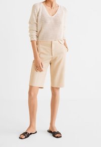 Beżowy sweter o dzianinowym wykończeniu z dekoltem w serek, w połączeniu z jasnobeżowymi dopasowanymi szortami. Model w czarnych klapkach, podkreślający swobodny, casualowy wygląd.