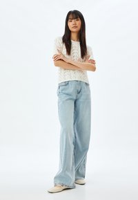 Gebreide, crèmekleurige trui met bloemmotieven, gecombineerd met lichtblauwe, wijde denim jeans. Model staat met de armen gekruist op een effen achtergrond.