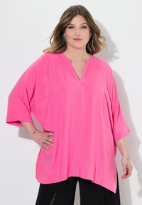 Blusa rosa, ampia e morbida, con scollatura a V e maniche corte. Presenta spacchi laterali e un orlo arricciato. Tessuto morbido e leggero.