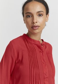 Blouse rouge à boutons avec un tissu texturé, présente des plis verticaux, un col mandarin et des détails de boutons à l'avant.