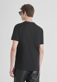 Camiseta de algodón negra con cuello redondo clásico y mangas cortas, con un pequeño detalle de logo en la parte posterior. Combinada con pantalones de cuero negros.